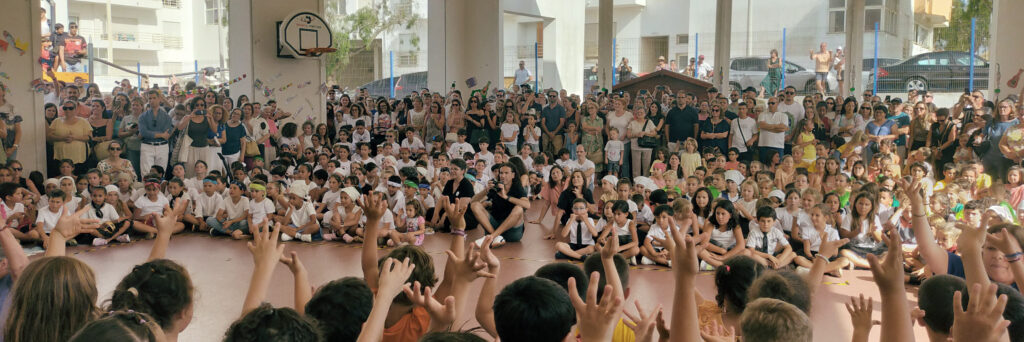 Um grande grupo de crianças senta-se de pernas cruzadas no chão de um ginásio, com as mãos erguidas, rodeado por uma multidão de adultos em pé, numa reunião escolar.